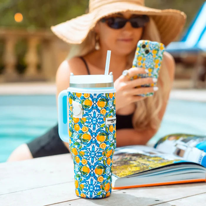 Yellow lemon-patterned tumbler carry case by Cocopup London on a table, woman by pool holding phone in background.