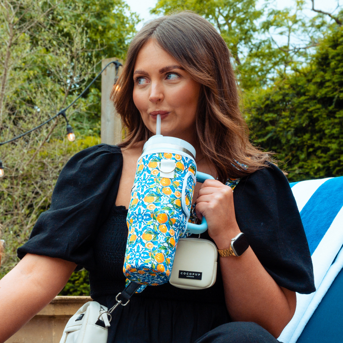Woman in a black dress holds a yellow lemon-patterned tumbler carry case by Cocopup London amid green outdoor foliage.
