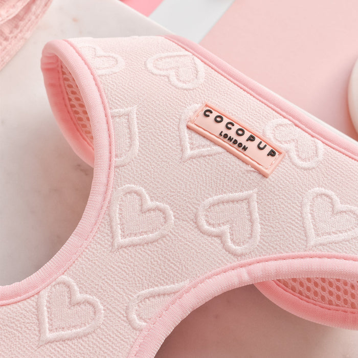 Close-up of the Luxe Baby Pink Heart Adjustable Neck Harness from Cocopup London, featuring an embossed heart pattern. The harness includes a tag with the text "COCOPUP LONDON" sewn onto it. Perfectly coordinating collars and leads are also available. The background showcases soft pink and white elements, enhancing the pastel theme.