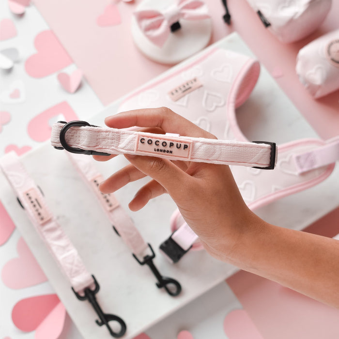 A hand holds the Luxe Baby Pink Heart Adjustable Neck Harness, Lead & Collar Bundle from Cocopup London, adorned with heart patterns and a tag reading "COCOPUP London." The background showcases matching leashes, dog leads, and accessories featuring the same heart design, set on a pink and white surface decorated with heart shapes.