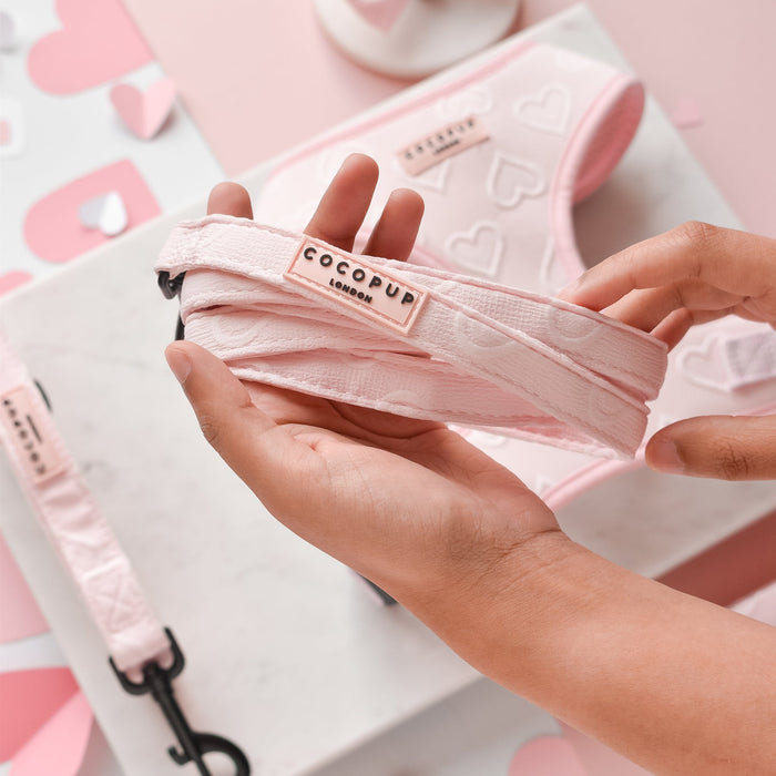 A person holds a pink dog lead from the "Cocopup London" brand. Other matching pet accessories, including the Luxe Baby Pink Heart adjustable neck harness and collar, are visible on a marble surface and pink background.