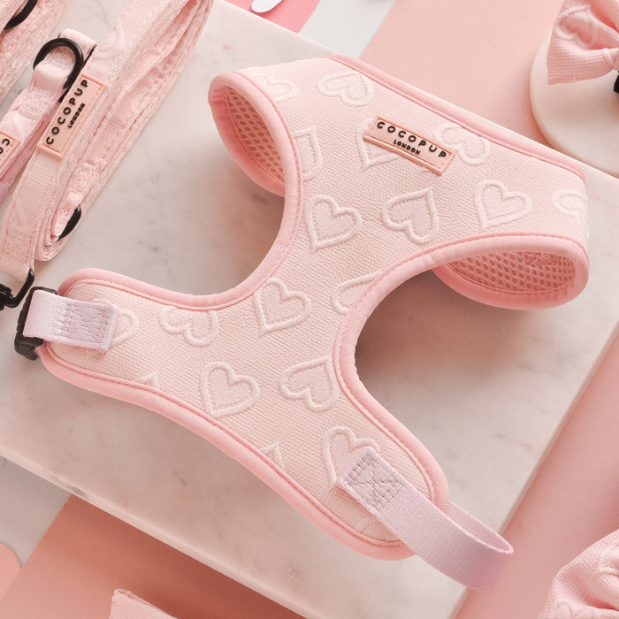 A Cocopup London Luxe Adjustable Neck Harness in Baby Pink Heart with delightful patterns is displayed on a marble surface. Surrounding it are matching accessories, including a leash, bows, and clips. The setting is adorned with cut-out heart shapes, enhancing the playful and loving theme.