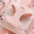 A Cocopup London Luxe Adjustable Neck Harness in Baby Pink Heart with delightful patterns is displayed on a marble surface. Surrounding it are matching accessories, including a leash, bows, and clips. The setting is adorned with cut-out heart shapes, enhancing the playful and loving theme.