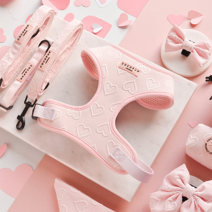 A collection of pink dog accessories from Cocopup London, featuring the Luxe Adjustable Neck Harness - Baby Pink Heart, a matching leash, bows, and bandanas, is beautifully arranged on a marble base. Small heart-shaped confetti and a light pink background enhance the charming and playful theme.