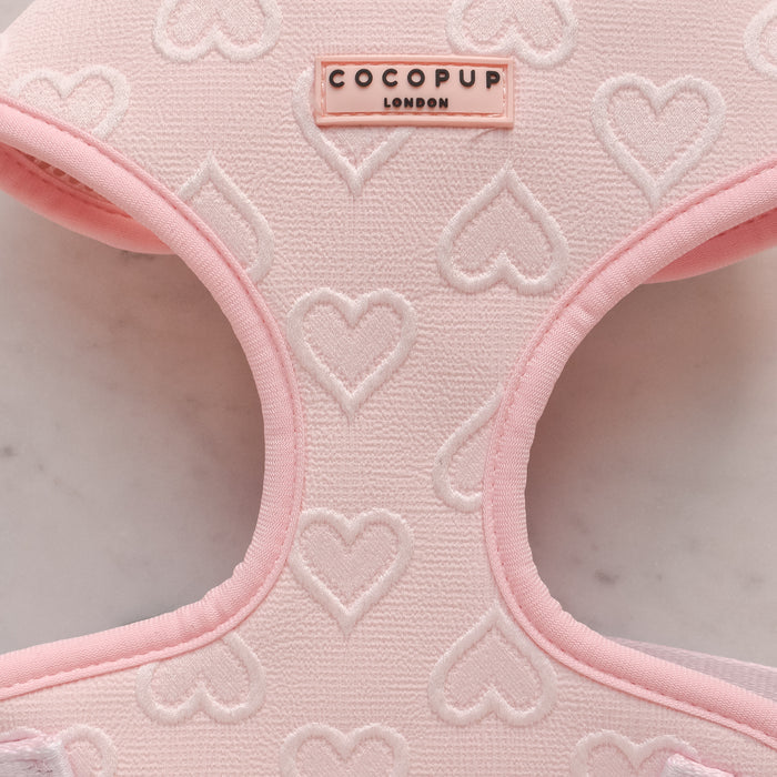 A Luxe Adjustable Neck Harness - Baby Pink Heart from Cocopup London is placed on a light marble surface. This dog harness, adorned with a pattern of white hearts, features an embroidered Cocopup London label at the top.