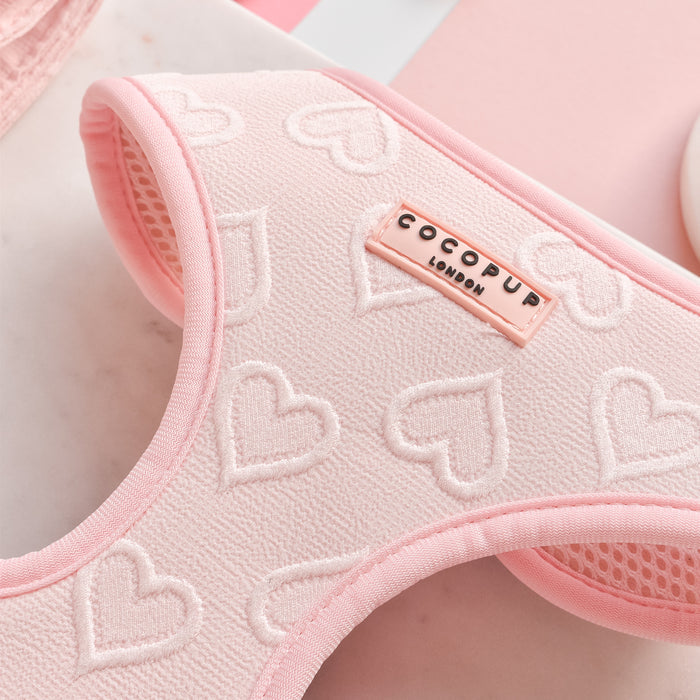Close-up image of the Luxe Adjustable Neck Harness in Baby Pink Heart by Cocopup London, featuring a pattern of embroidered hearts. The harness includes a stitched label that reads "COCOPUP LONDON." The background showcases shades of pink and white, contributing to a soft, playful aesthetic.