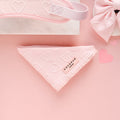 The Luxe Bandana - Baby Pink Heart by Cocopup London, featuring embossed heart patterns, is centered on a light pink surface. It is surrounded by other pink accessories, including a sleep mask and a hair bow, both similarly patterned with hearts. Nearby lie two small pink heart cutouts.