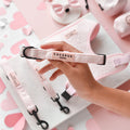 Hand holding a pink heart-patterned Cocopup London Luxe collar, surrounded by matching pink pet accessories.
