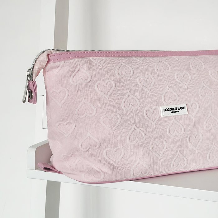 Baby pink expandable makeup bag with textured hearts and a silver zipper, on a white shelf.