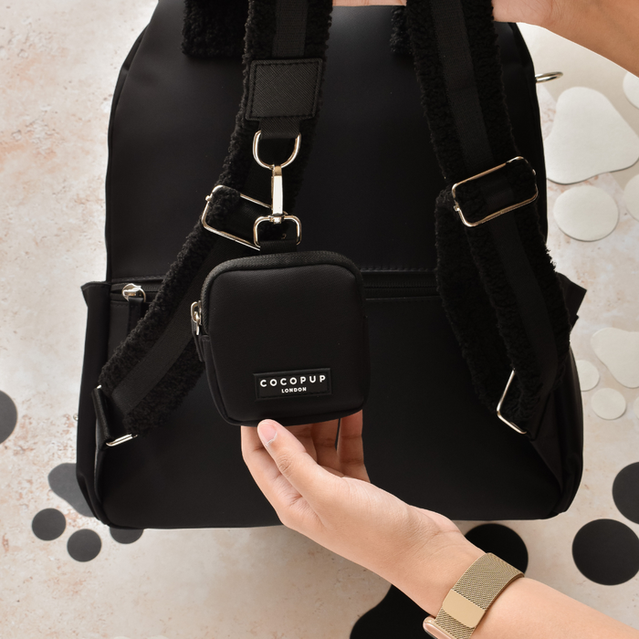 A hand clips a beige Cocopup London dog walking bag to the straps of a black rucksack.