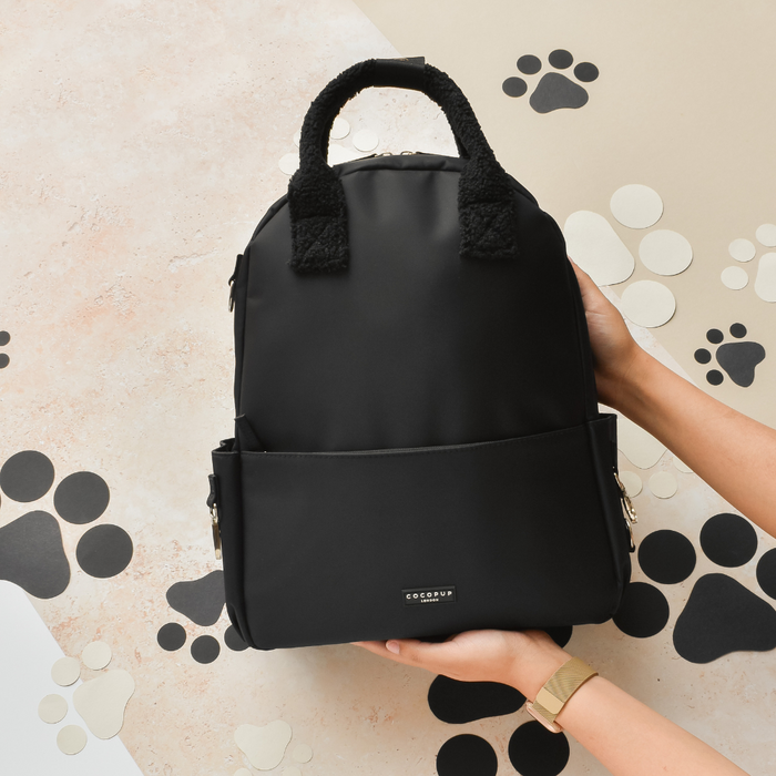 Black Cocopup London Rucksack with a fuzzy handle, held in hands and surrounded by black and white paw print designs.