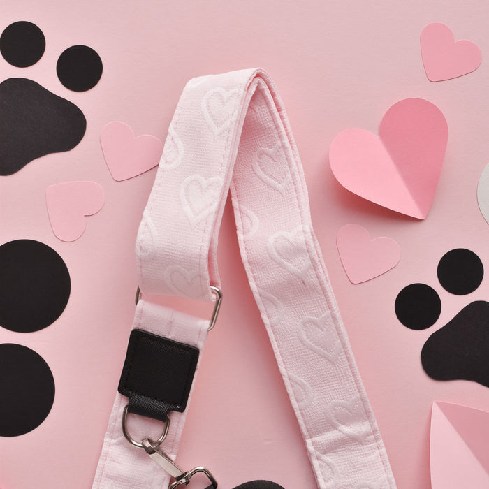 A pink dog leash with heart patterns is placed on a pink background. Surrounding the leash are heart-shaped and black paw print cutouts, arranged in a playful and decorative manner. The scene is complemented by a Luxe Bag Strap - Baby Pink Heart from Cocopup London, adding charm to the display.