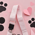 A pink dog leash with heart patterns is placed on a pink background. Surrounding the leash are heart-shaped and black paw print cutouts, arranged in a playful and decorative manner. The scene is complemented by a Luxe Bag Strap - Baby Pink Heart from Cocopup London, adding charm to the display.