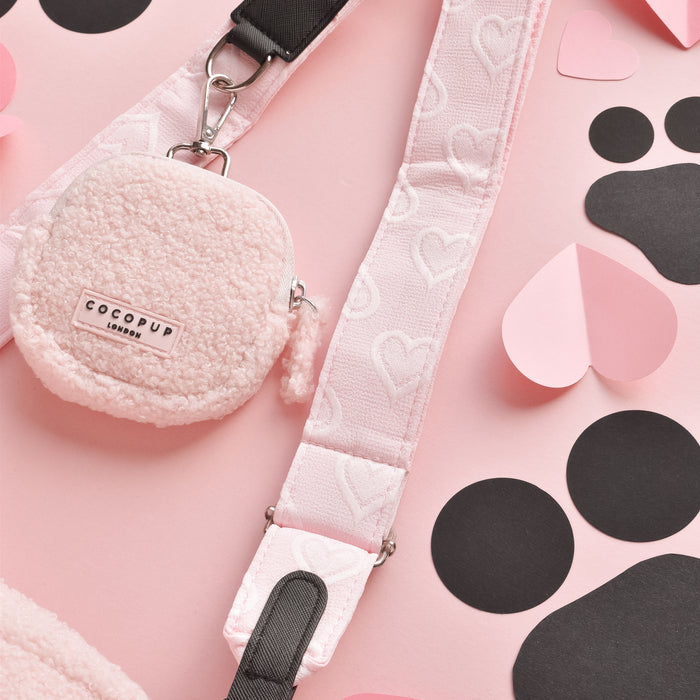 A pink crossbody bag with a soft, fluffy texture and the Luxe Bag Strap - Baby Pink Heart by Cocopup London is laid on a pastel pink surface. Surrounding it are black and pink paper cutouts in shapes like hearts, circles, and paw print designs, giving it an adorable Cocopup Dog Walking Bag vibe.