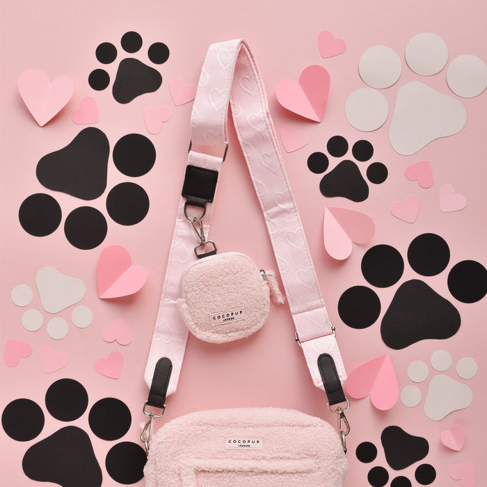 A Cocopup London Dog Walking Bag with a matching round pouch and Luxe Bag Strap in Baby Pink Heart is laid on a pink background adorned with black paw prints and various pastel-colored 3D heart cutouts. The bag, featuring small hooks for the strap and a fluffy, textured exterior, creates the perfect accessory set.