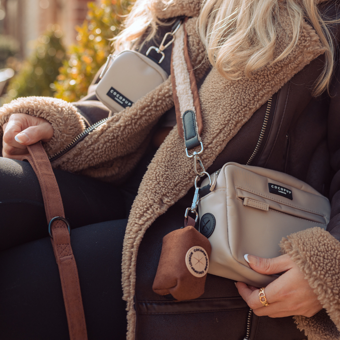 A person wearing a brown shearling jacket holds the strap of their Dog Walking Bag. The beige crossbody bag, enhanced with a small accessory and keychain, pairs perfectly with their black leggings and ring. Sunlight highlights the scene, adding warmth and charm to the moment. They're using the Bag Strap - Brown Aviator from Cocopup London.