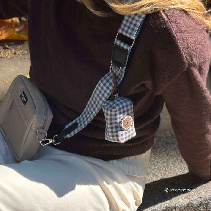 A close-up lifestyle shot of a person wearing a brown knit sweater and cream pants, carrying a small beige crossbody bag and a matching blue-and-brown gingham poop bag holder