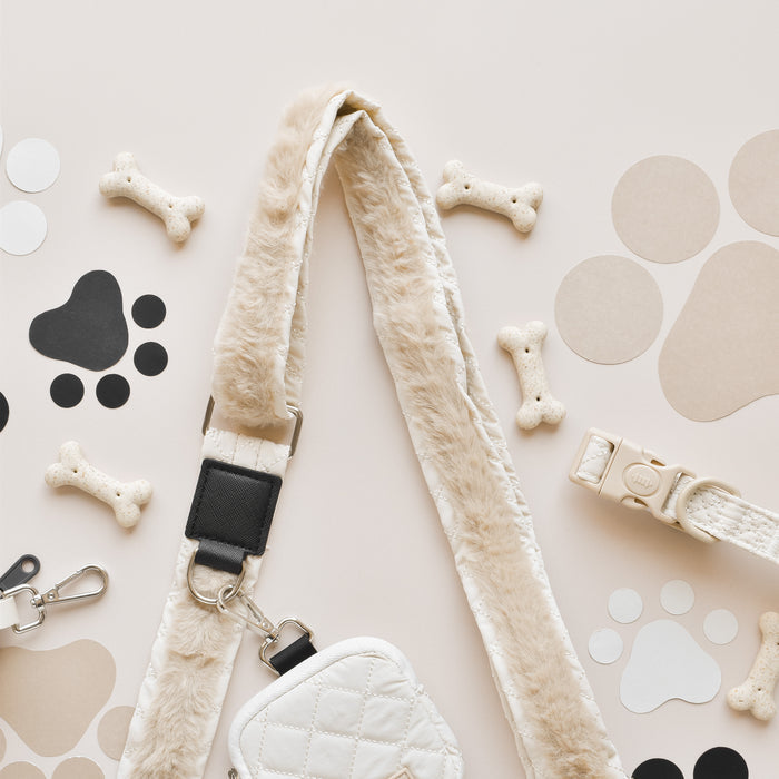 A flat lay image displays a collection of dog accessories, including a fur-lined leash, a small quilted dog walking bag from Cocopup London with its matching Bag Strap - Quilted, and scattered bone-shaped dog treats. Various black and beige paw print cutouts are also arranged around the items on a light background.