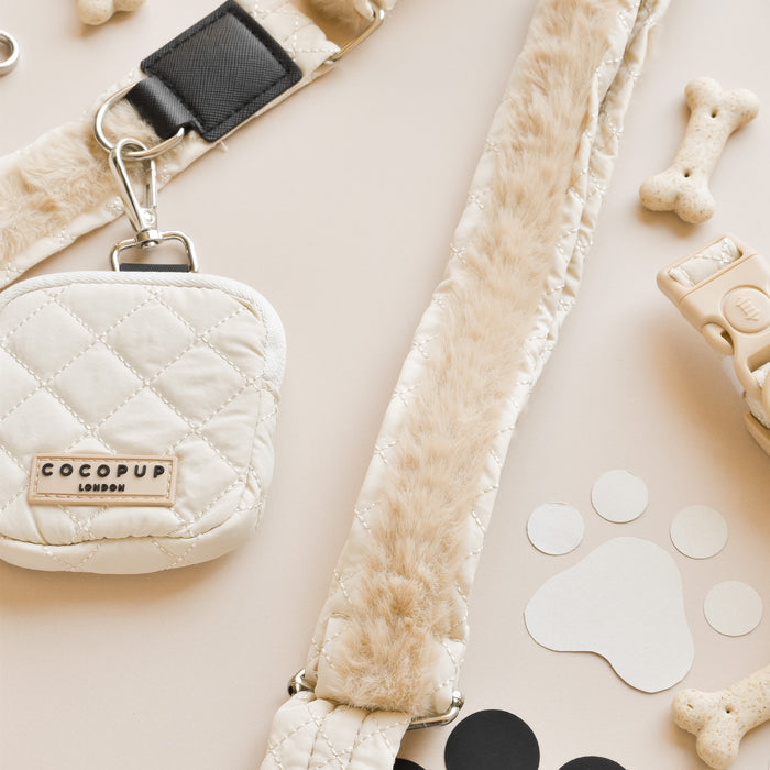 A close-up of beige quilted dog leash accessories, featuring a Cocopup London small pouch attached to a Cocopup London Bag Strap - Quilted. The setup includes a section with fur trim, bone-shaped biscuits, a beige waste bag holder, and paw-print stickers on a cream-colored surface. Perfect for stylish dog walks!