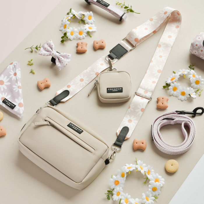 A stylish tan crossbody bag and small pouch with Cocopup London's matching Bag Strap - Daisy Chain are displayed on a beige background. The scene also features dog accessories, including a leash, a poop bag holder, fabric daisies, treats, and a patterned bow and bandana. Perfect for dog walks, this chic ensemble is the ultimate Dog Walking Bag set.