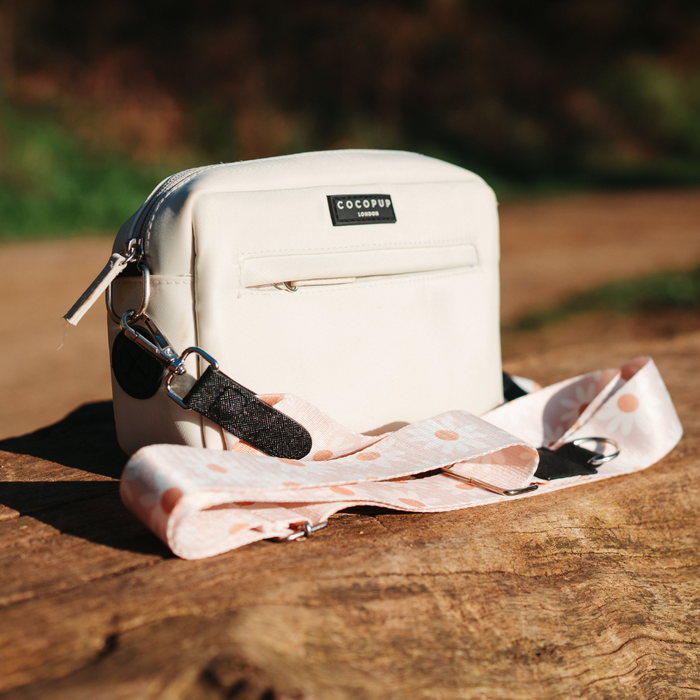 A small, white crossbody dog walking bag with a black logo tag that reads "Cocopup London" sits on a wooden surface. The bag features a front zipper pocket and is accompanied by the Bag Strap - Daisy Chain, a light pink strap with white polka dots. The outdoor background is slightly blurred.