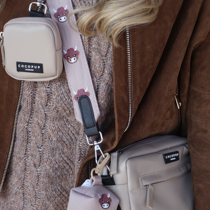 Person in brown jacket holding beige Cocopup London bags with a tan and cream Highland Cow print bag strap.