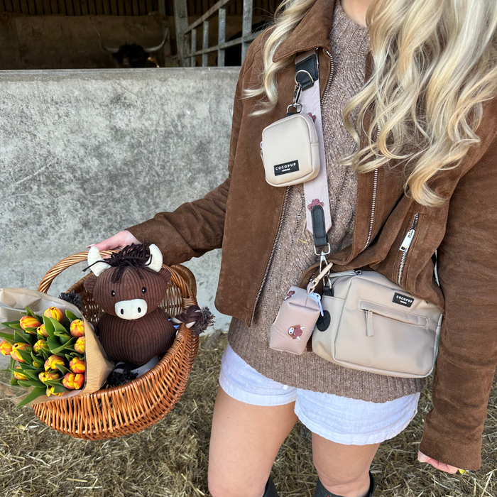 Woman in a brown jacket holds a wicker basket with tulips and a plush Highland Cow poop bag holder attached.