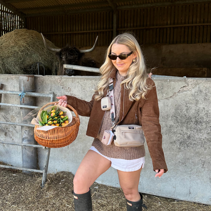 Woman in brown jacket with sunglasses holds yellow tulips, wearing a tan and white cow print Cocopup London bag strap.