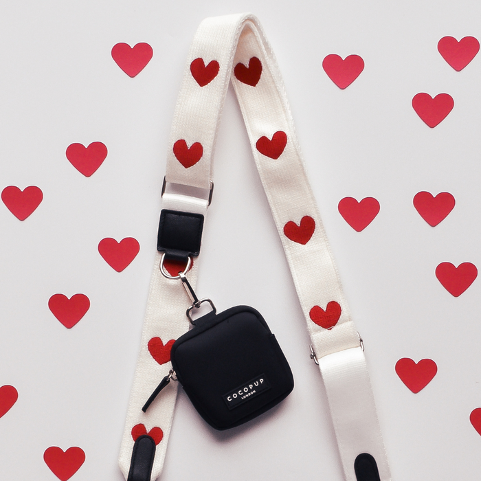 White Cocopup London bag strap with red hearts and black case on a background of scattered red heart shapes.