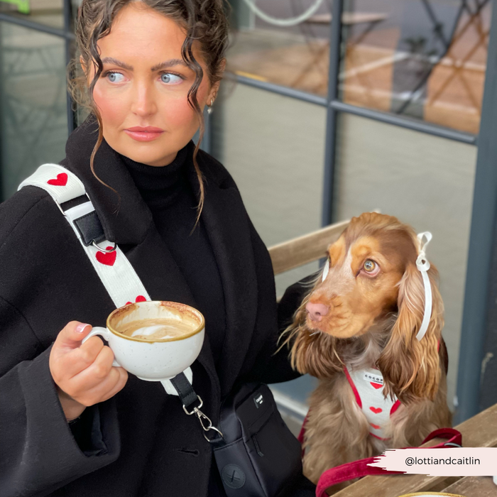 Woman with curly hair holds coffee; her dog wears a pink knitted Cocopup London bag strap with heart pattern.