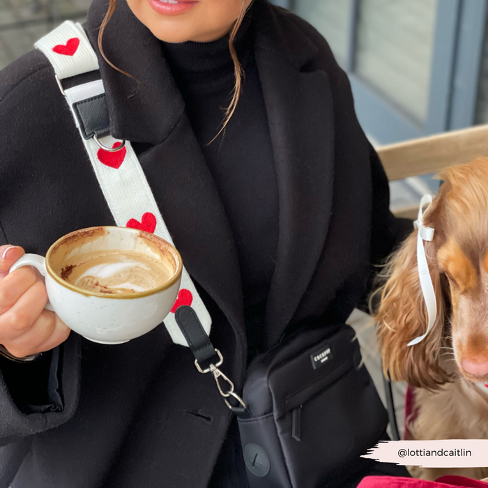 Woman in a black coat holds cappuccino, wearing a Cocopup London bag strap with hugs pattern; dog has Valentine’s accessory.