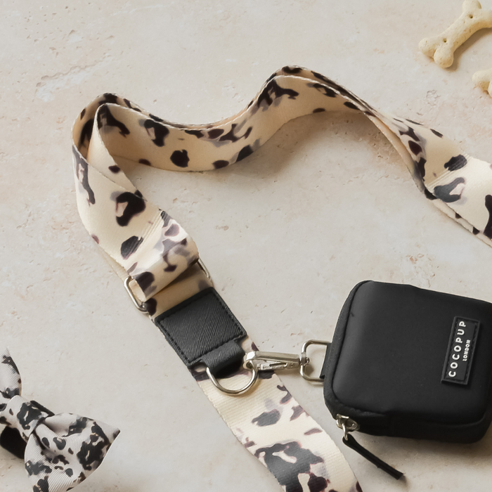 A beige and black leopard print lanyard with a black rectangular pouch from the Cocopup London Dog Walking Bag collection is clipped onto a silver hook, resting on a light-colored surface. Nearby lies a tied fabric bow featuring a similar leopard print, while parts of an ivory tort bone-shaped item called the Bag Strap - Ivory Tort are visible.