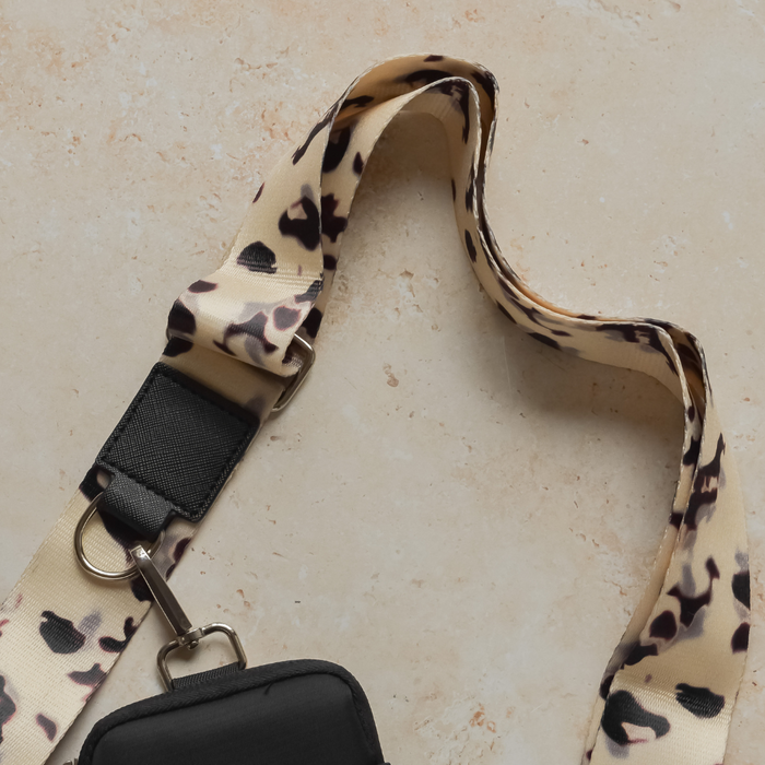 The Bag Strap - Ivory Tort from Cocopup London, featuring a beige and black leopard print pattern, an adjustable length, and a black attachment clip, is displayed on a beige, textured surface. The strap is designed for the Ivory Tort Cocopup Dog Walking Bag and is arranged loosely in a curved shape, ready to attach to a small bag or pouch.