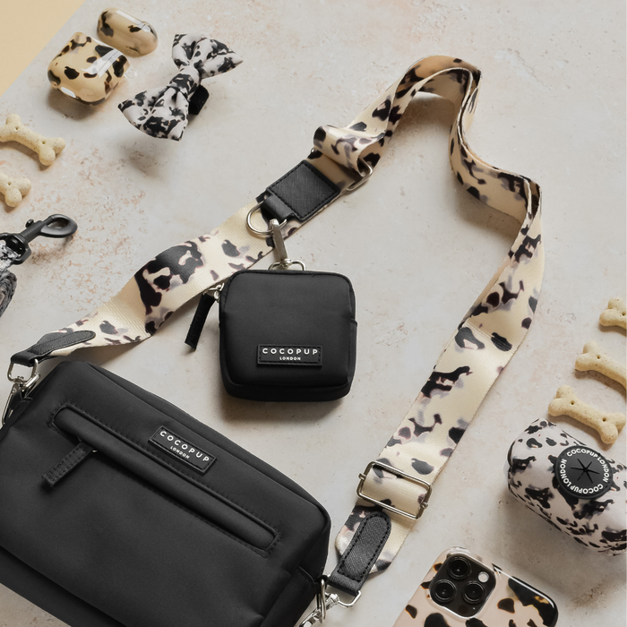 A black Cocopup Dog Walking Bag with the "Cocopup London" label, featuring an adjustable length detachable strap in a chic Ivory Tort pattern. Surrounding items include a matching small pouch, dog waste bag holder, bow tie, phone case, lip balm, and scattered dog bone treats.