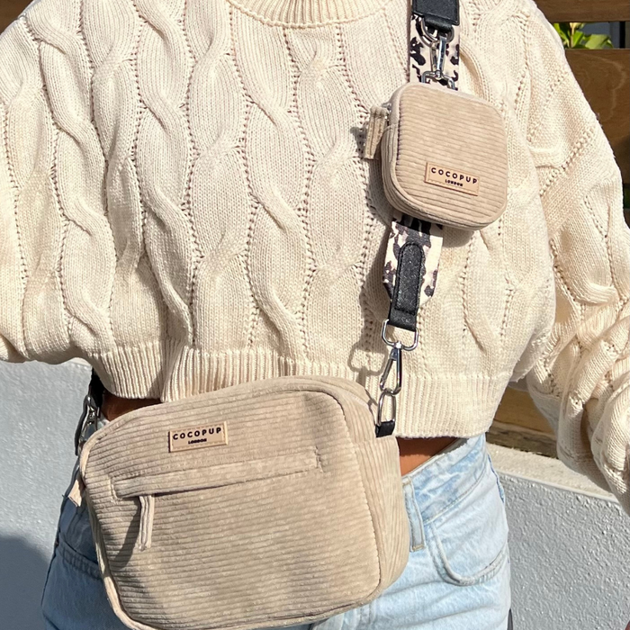 A person wearing a cream-colored cable knit sweater and light blue jeans is shown. They are carrying a beige Cocopup London Dog Walking Bag with an adjustable Ivory Tort strap that has the brand name "Cocopup London" on it. The bag features a smaller matching pouch attached to the strap, which displays a black-and-white animal print.