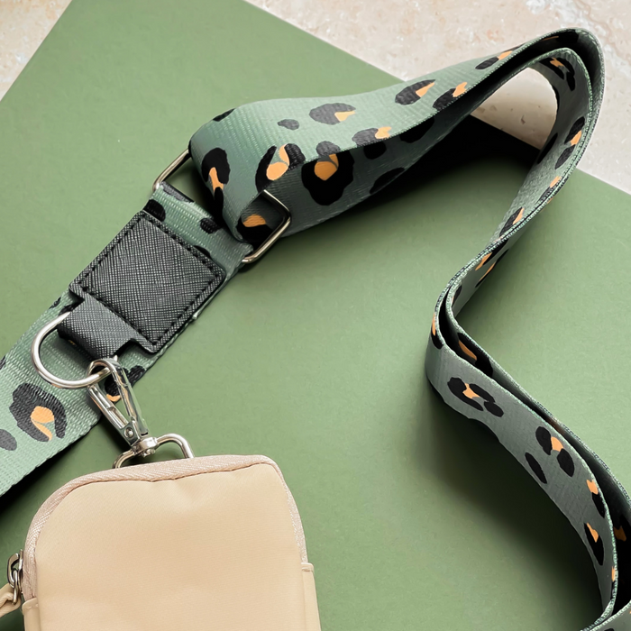 The Cocopup London Bag Strap - Khaki Leopard features a beige bag with a clip attached to a green shoulder strap adorned with a striking black and orange leopard print pattern. Displayed on a green surface, this stylish piece is part of the Khaki Leopard Collection.
