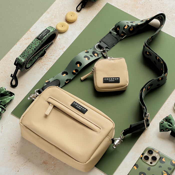A stylish flat lay features a beige crossbody bag and matching small pouch accessorized with Cocopup London’s Bag Strap - Khaki Leopard. Surrounding the Cocopup Dog Walking Bag are a dog leash, collar, bow tie, cookies, and a smartphone with a green leopard print case, all laid out on a green-and-cream surface.