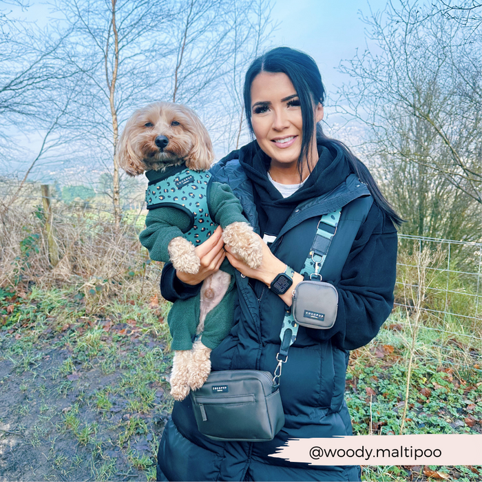 A woman with long dark hair stands outdoors holding a small dog. She is dressed in a black jacket, and the dog is wearing an outfit from the Khaki Leopard Collection. They are in a natural setting with bare trees and a fence in the background, and you can spot her Cocopup London Bag Strap in Khaki Leopard. The Instagram handle @woody.maltipoo is displayed.