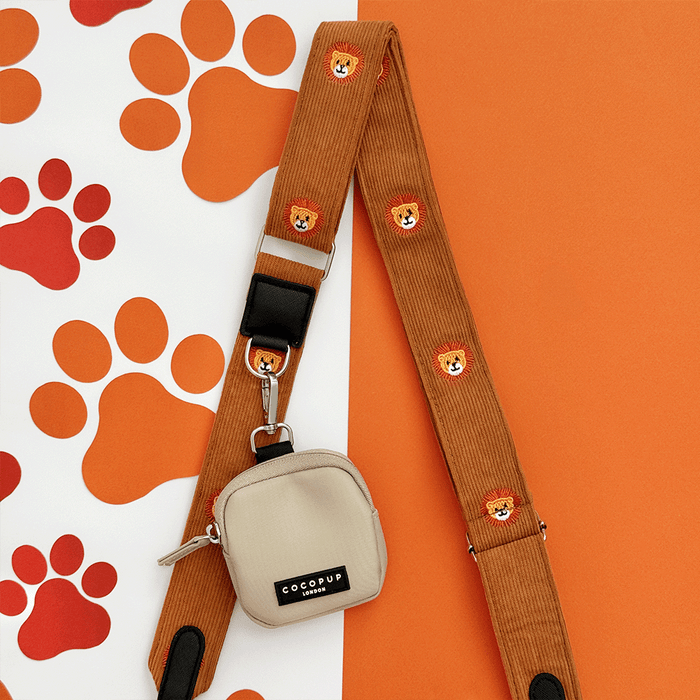 A brown / orange corduroy bag strap, with embroidered lion faces. Attached is a small beige pouch for dog treats.