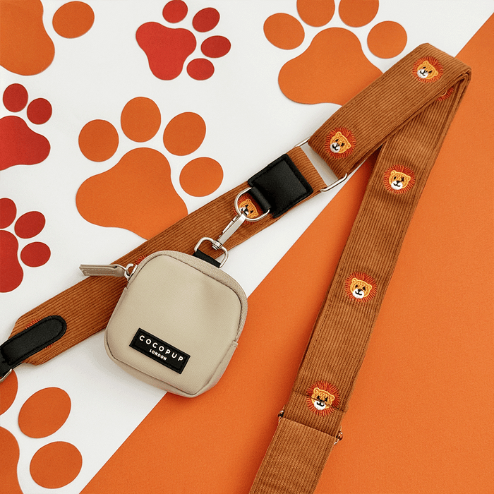 A brown / orange corduroy bag strap, with embroidered lion faces. Attached is a small beige pouch for dog treats.