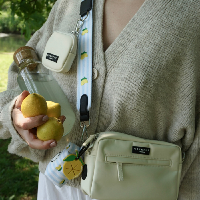 Person holding lemons and a glass bottle, wearing a cream cardigan and lemon-patterned fabric bag strap.