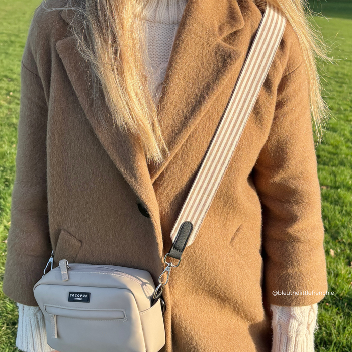 Woman in a tan coat carrying a Cocopup London bag with a linen-striped strap, standing on green grass.