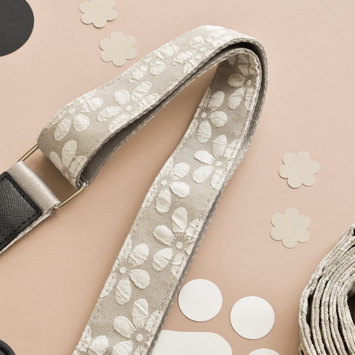 A beige camera strap featuring a white floral pattern is laid on a light pink surface. Surrounding the strap are various beige and white circular and flower-shaped paper cutouts, resembling those adorning the Luxe Bag Strap - Mocha Flower by Cocopup London, making it a stylish accessory. The background includes subtle black elements for contrast.