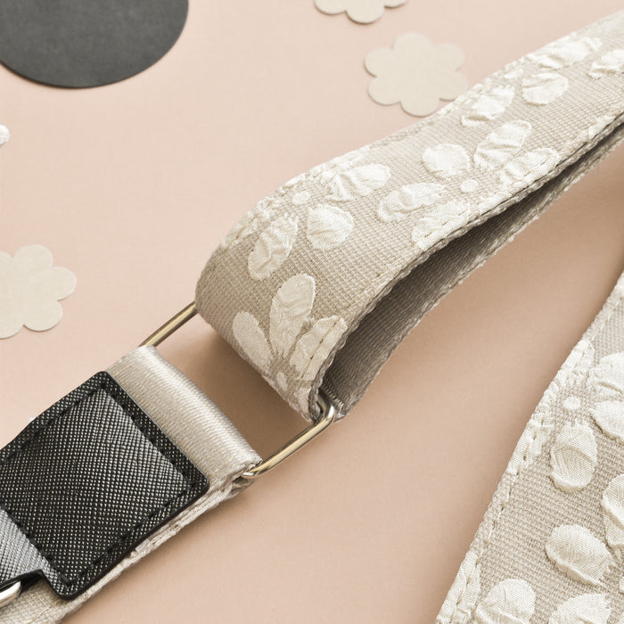 A close-up shot reveals a fabric strap adorned with a floral pattern in light beige and white colors. The Luxe Bag Strap - Mocha Flower by Cocopup London is partially attached to a metallic ring. In the background, cutouts of flowers and circles with neutral prints are scattered across a neutral-toned surface.