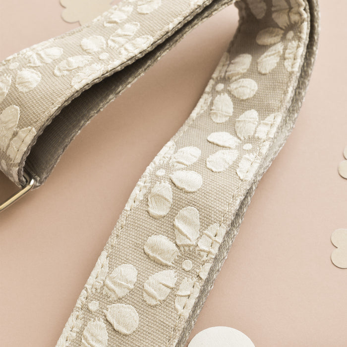 A close-up of a beige fabric strap with an embossed floral pattern, perfect as the Luxe Bag Strap - Mocha Flower by Cocopup London. The accessory lies on a light peach background, adorned with small circular paper cutouts in matching hues.