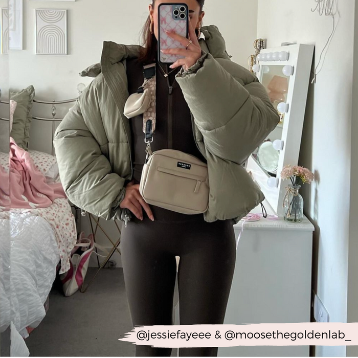 A person is taking a mirror selfie in a bedroom. They are wearing a green puffer jacket, brown bodysuit, and carrying two cream-colored bags across their chest with Cocopup London Luxe Bag Strap - Mocha Flower. The room has a made bed, a vanity with lights and flowers, and artworks on the wall.