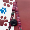 Maroon plaid dog leash with black pouch beside matching Mulberry Plaid bag strap on paw print fabric.