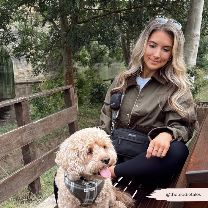Woman sitting by her tan dog in a plaid harness with an olive check dog walking bag bundle, outdoors near trees and water.