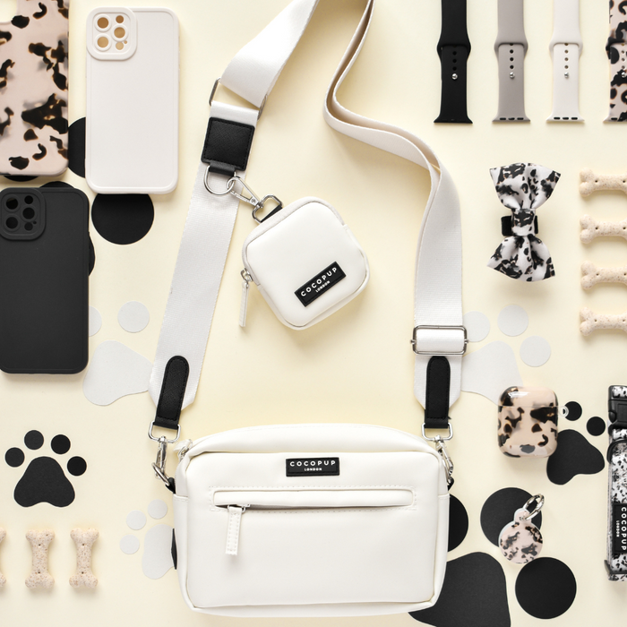 A flat lay image showcases a variety of black, white, and patterned accessories. At the center is a Bag Strap - Oyster White by Cocopup London, featuring a small pouch attached. Surrounding the strap are phone cases, polyester wearable bands, bow ties, and Cocopup dog bone treats in coordinated colors and designs.