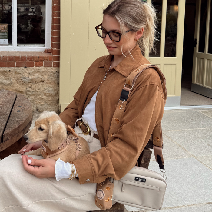 Woman in brown jacket seated outdoors with beige puppy on her lap, beige Cocopup bag strap with paw print pattern beside her.